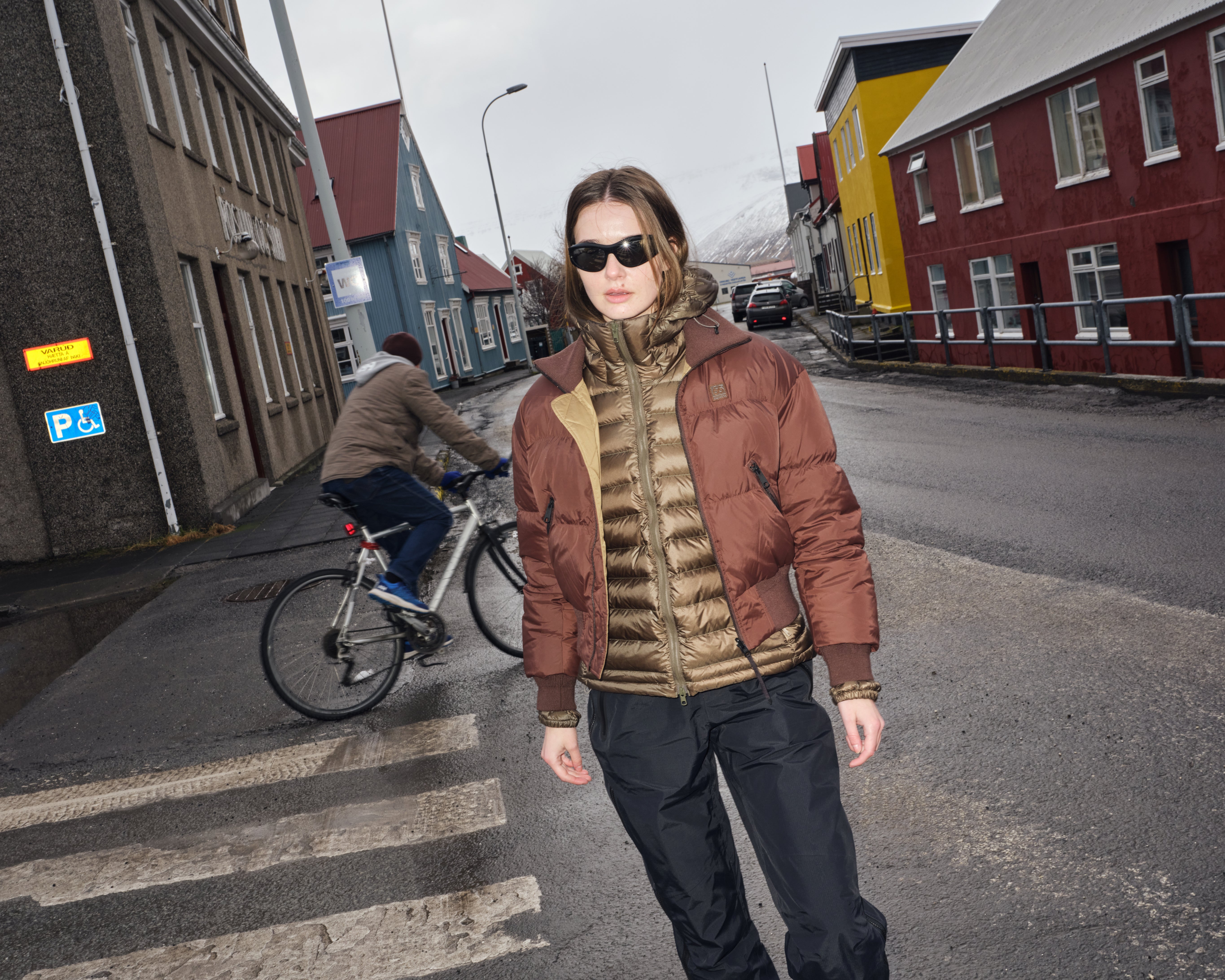 Person wearing sunglasses and a brown jacket standing on a street with colorful buildings in the background.