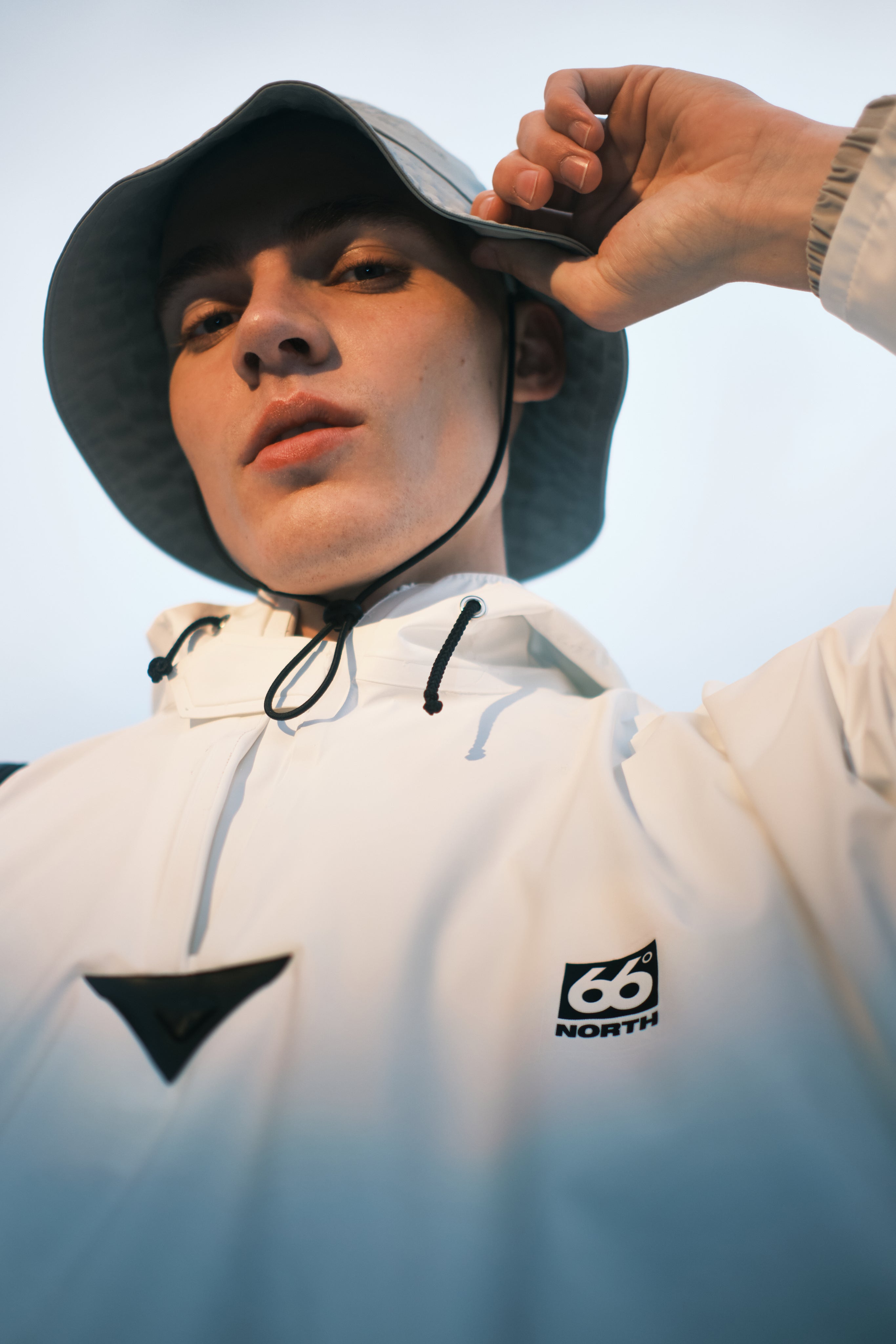 Person wearing a white hoodie with a logo, adjusting a black hat against a clear sky.