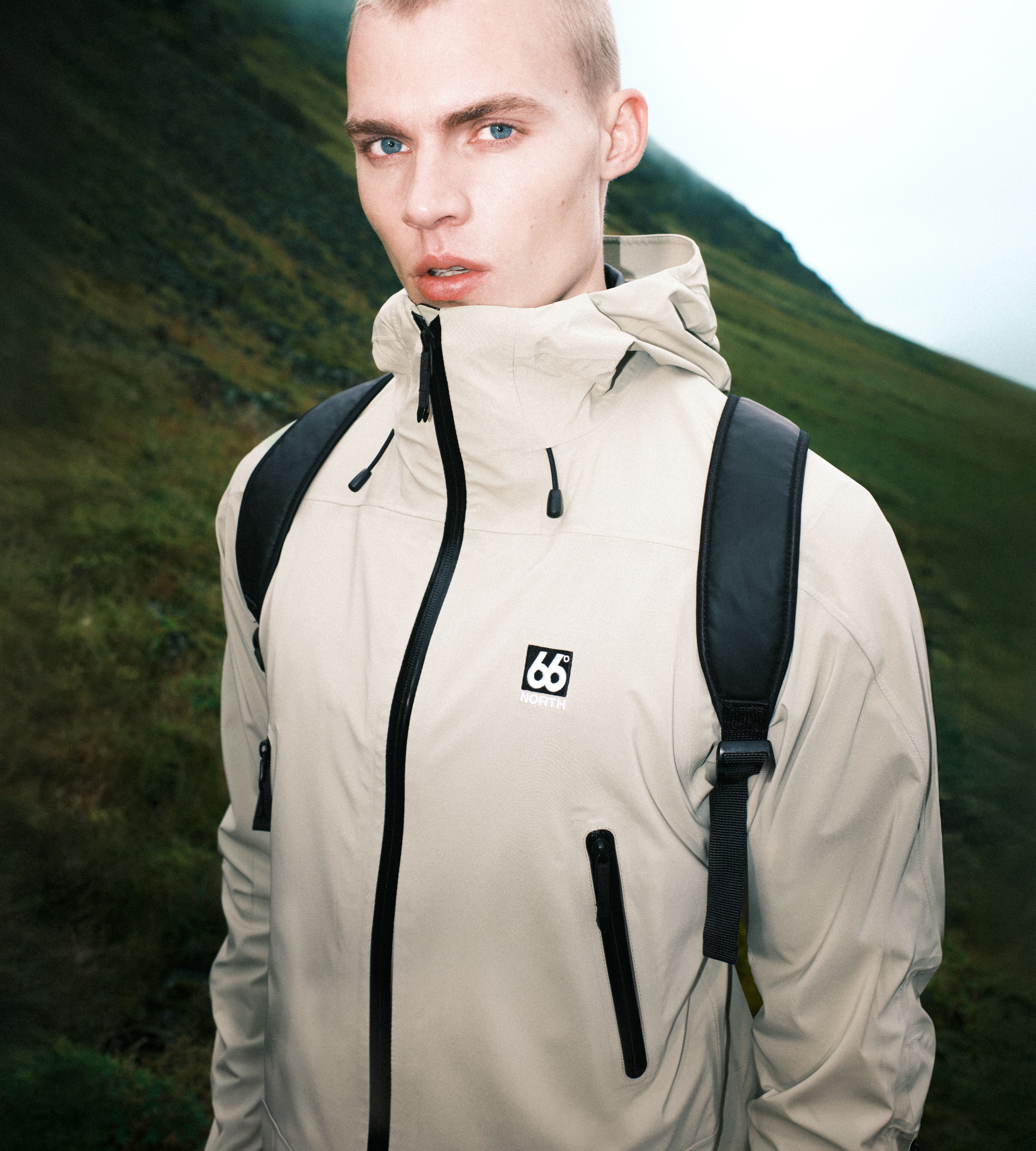 Person wearing a beige jacket with a logo, standing in a mountainous landscape.