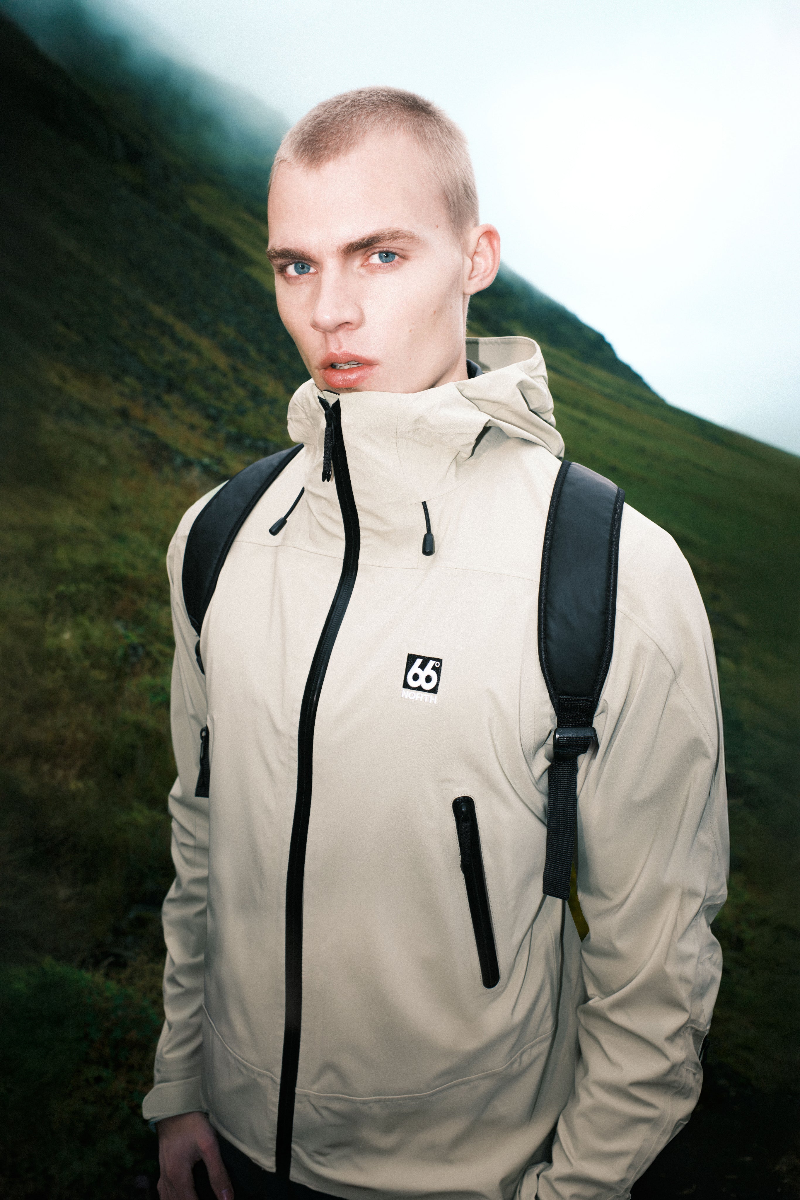 Person wearing a beige jacket with a logo, standing in a mountainous landscape.