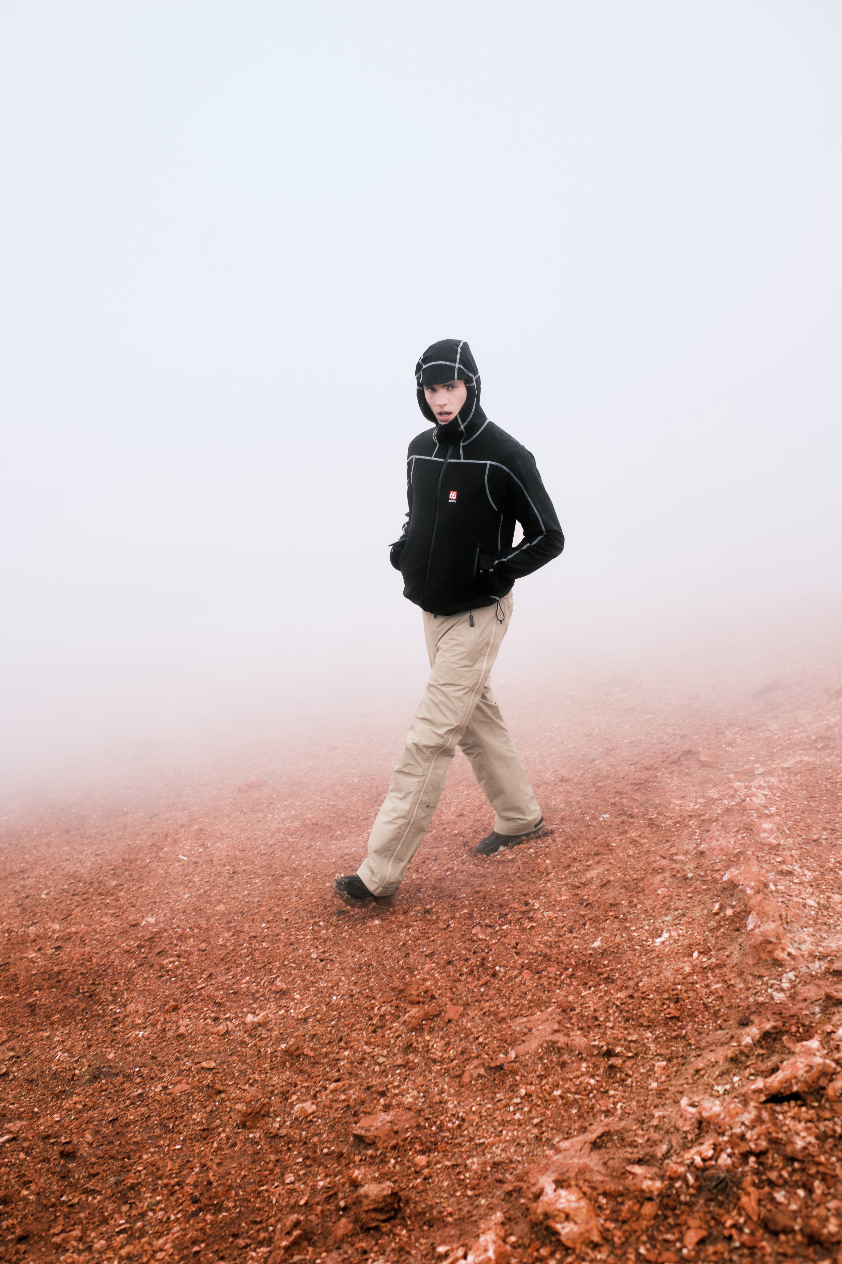 Person wearing a black jacket and beige pants walking on a red-brown surface with a white background
